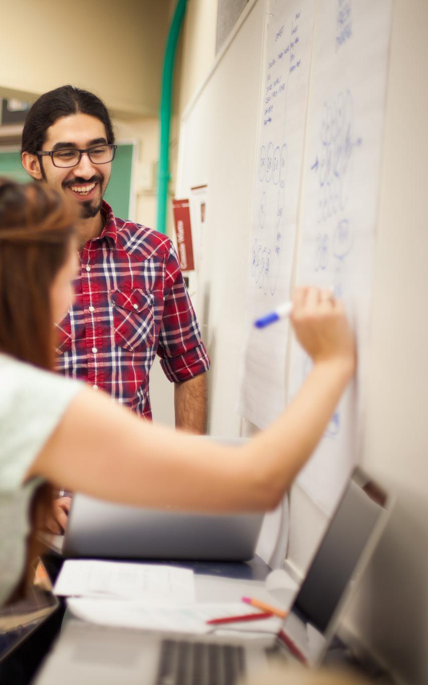 a group of five smiling students in casual clothing facing a whiteboard with one student drawing on the board with a blue marker and a laptop in the foreground